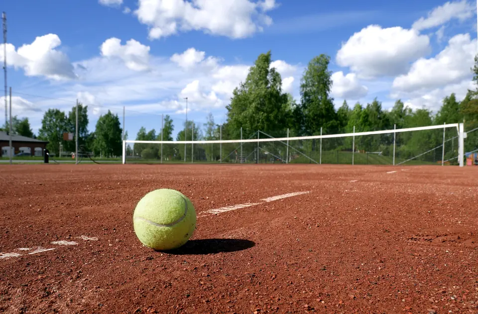 En tennisball på en grusbane med nett og grønne løvtrær i bakgrunnen