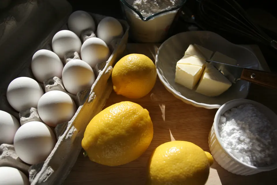 Bakeingredienser i sollys: egg i eggkartong, sitroner, smør i skål og melis på et trebrett