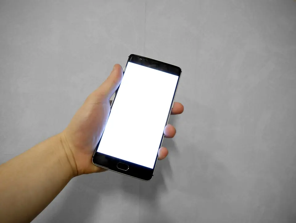 Hånd som holder en smarttelefon med hvit, tom skjerm foran en grå vegg