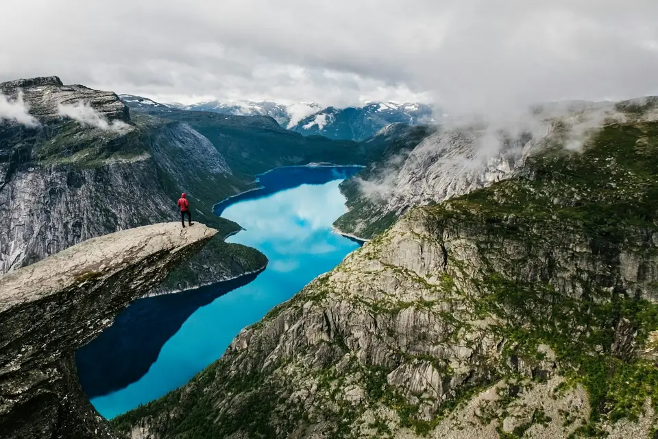 Person i rød jakke står ytterst på Trolltunga høyt over en turkis innsjø mellom fjell