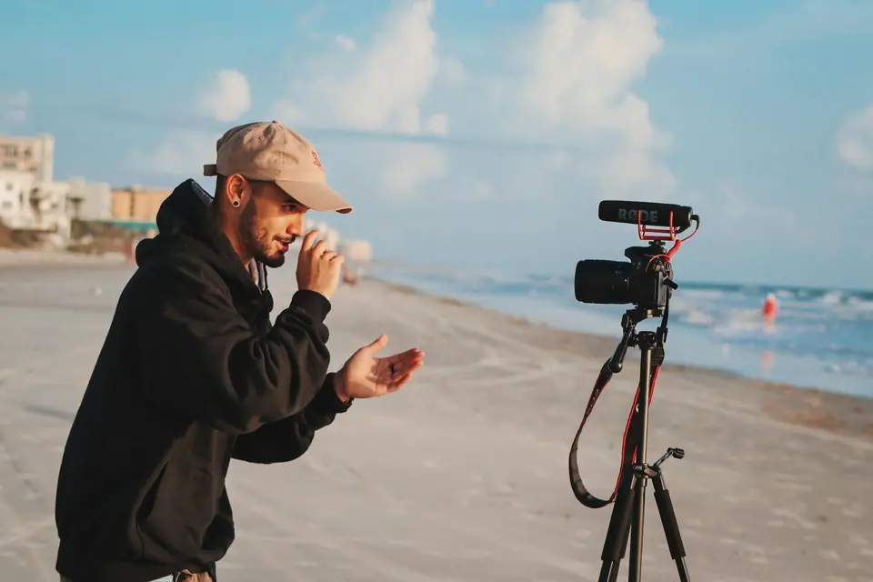Mann med caps snakker til et kamera på stativ på en strand