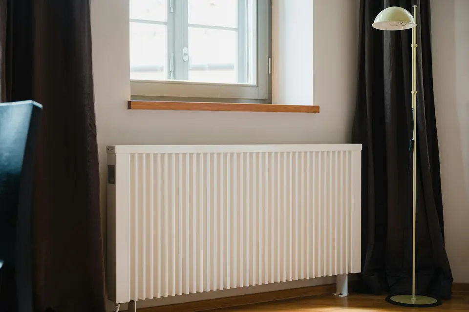 Hvit elektrisk panelovn under et vindu i en stue med mørke gardiner
