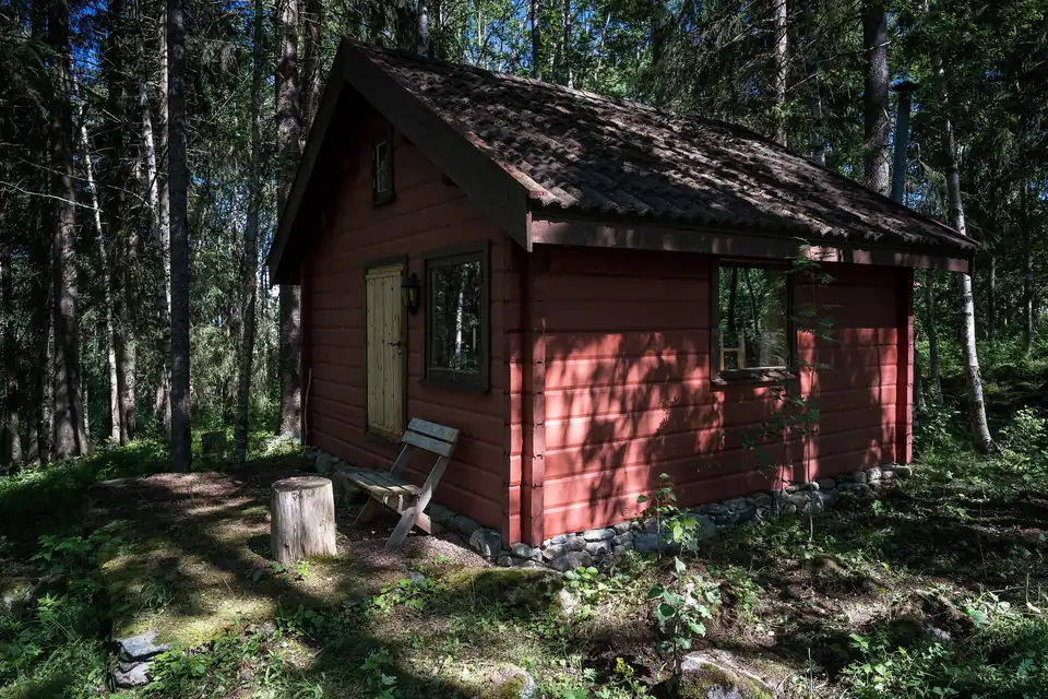 Liten rød tømmerhytte med benk og stubbe i en solfylt granskog