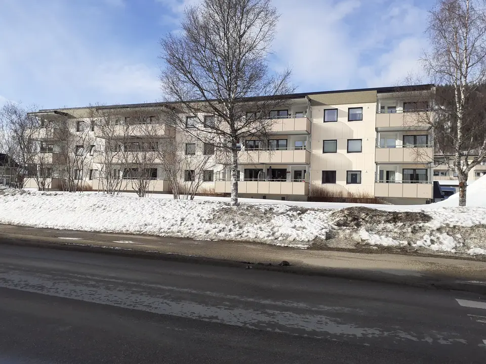 Lys boligblokk i tre etasjer med balkonger bak bjørketrær og snø i Mo i Rana
